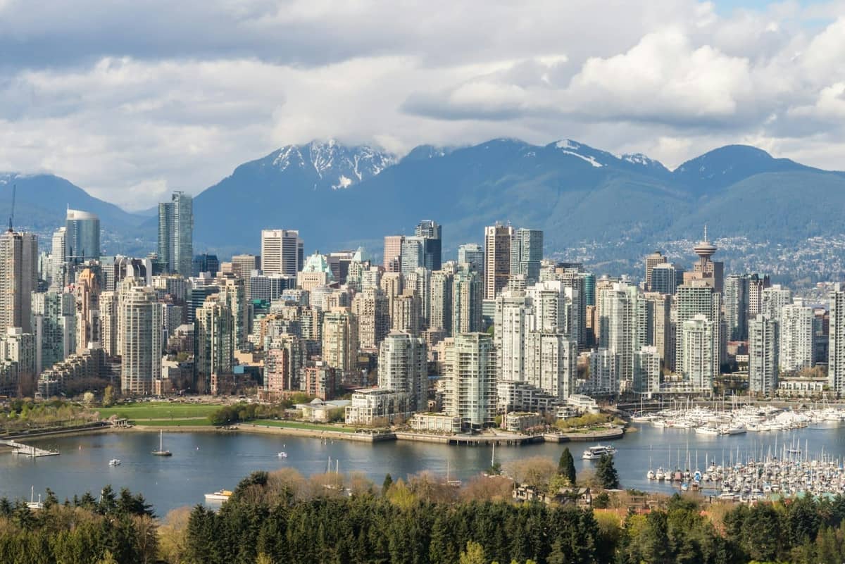 Skyline von Vancouver mit modernen Gebäuden, Wasserweg, Marina, üppigen Bäumen und schneebedeckten Bergen unter einem Mix aus Wolken und Sonne.