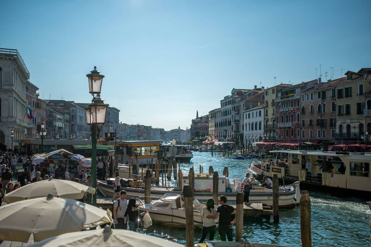 Belebte Szene am Canal Grande in Venedig an einem strahlenden Sommertag mit Menschenmassen, Booten, Sonnenschirmen und historischen Gebäuden unter klarem blauem Himmel.