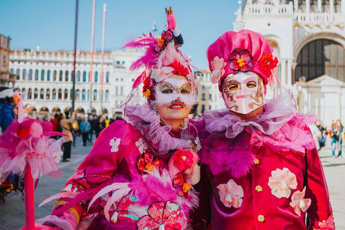 Zwei Personen in aufwendigen rosafarbenen venezianischen Karnevalskostümen und Masken posieren bei hellem Tageslicht auf der Piazza San Marco mit historischen Gebäuden im Hintergrund.