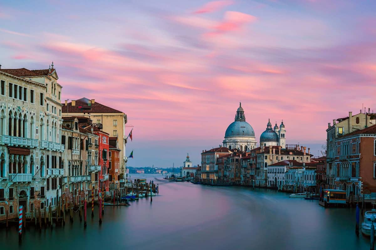 Venedigs Canal Grande bei Sonnenuntergang mit der Basilika Santa Maria della Salute im Hintergrund, flankiert von historischen Gebäuden und zartrosa Wolken am Himmel.