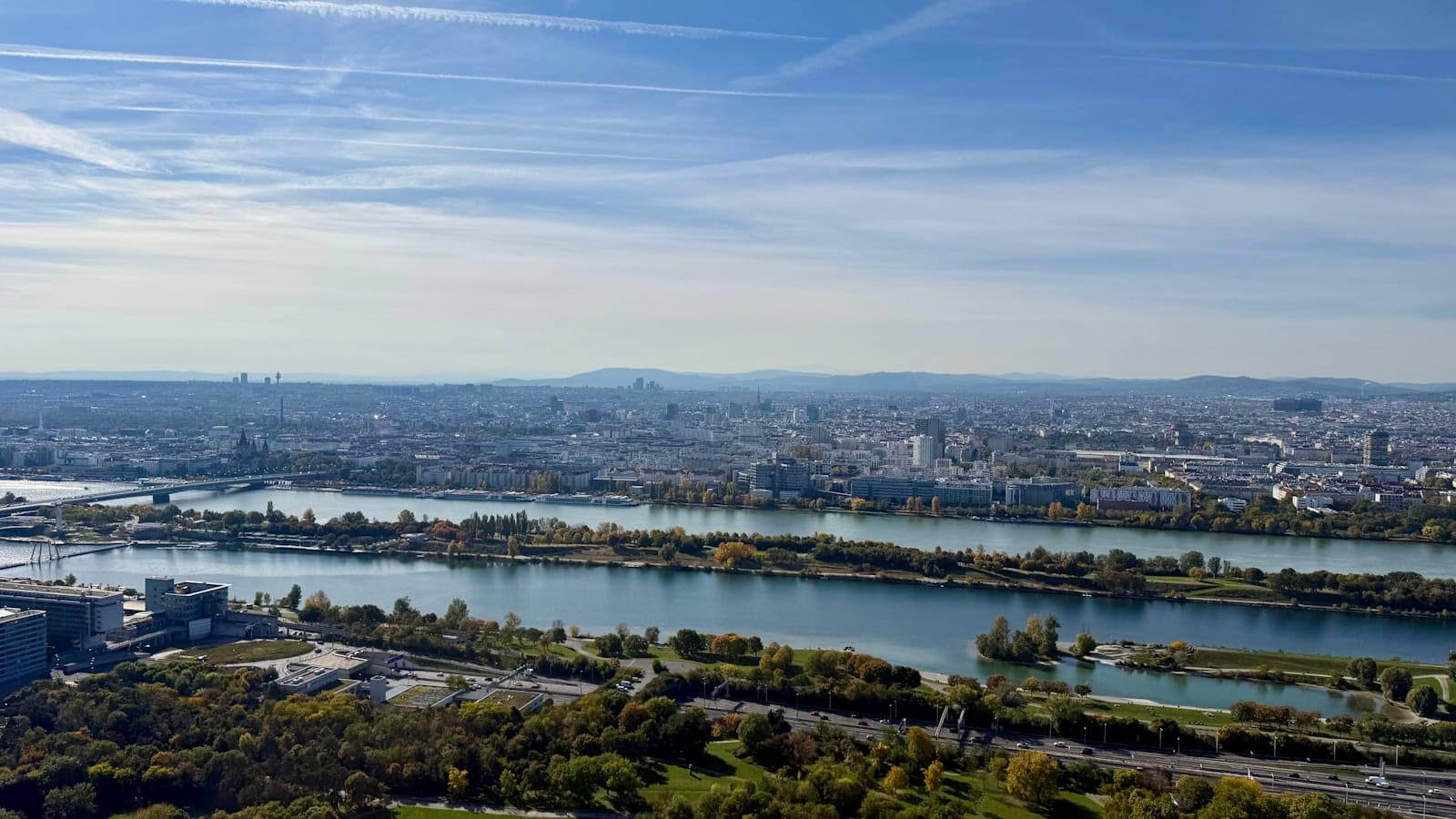 Ampia vista aerea di Vienna con il fiume Danubio, il paesaggio urbano e le montagne lontane sotto un cielo blu parzialmente nuvoloso