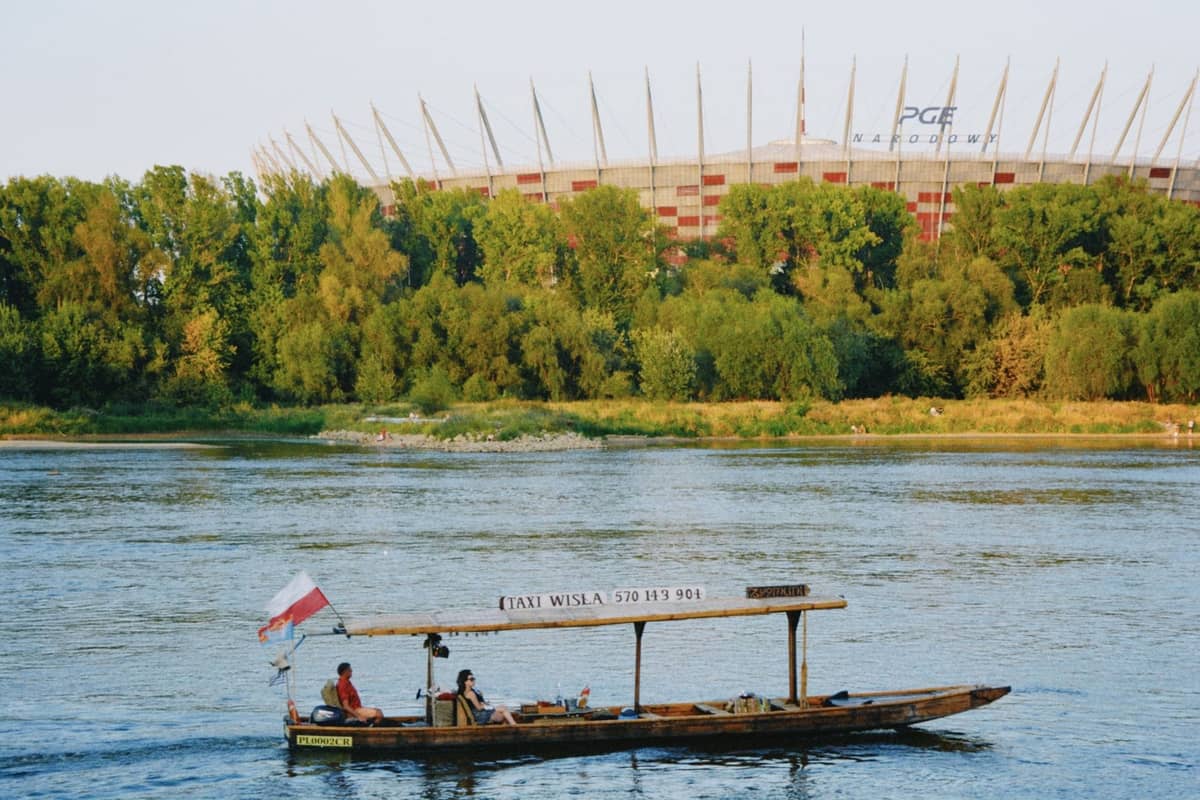 Tradycyjna drewniana łódź z ludźmi pływającymi po Wiśle, w tle stadion PGE Narodowy w Warszawie i bujne letnie drzewa.