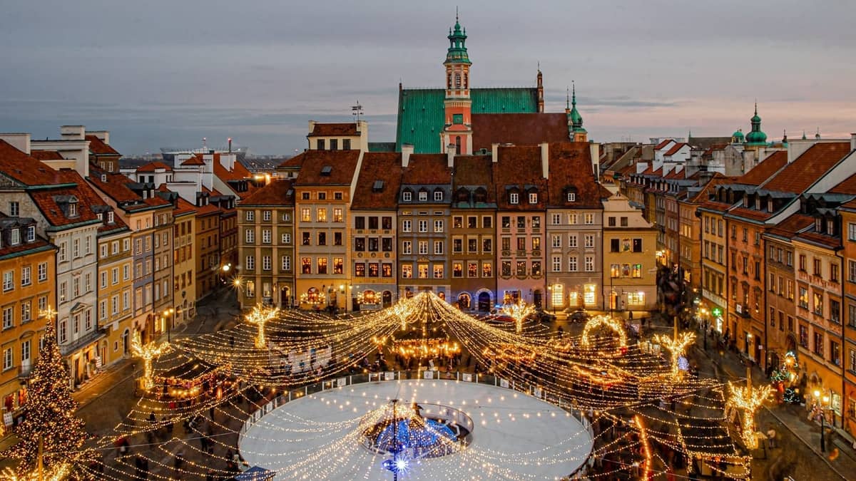 Rynek Starego Miasta w Warszawie zimą z świątecznymi lampkami bożonarodzeniowymi, lodowiskiem, kolorowymi historycznymi kamienicami i dużym kościołem w tle.