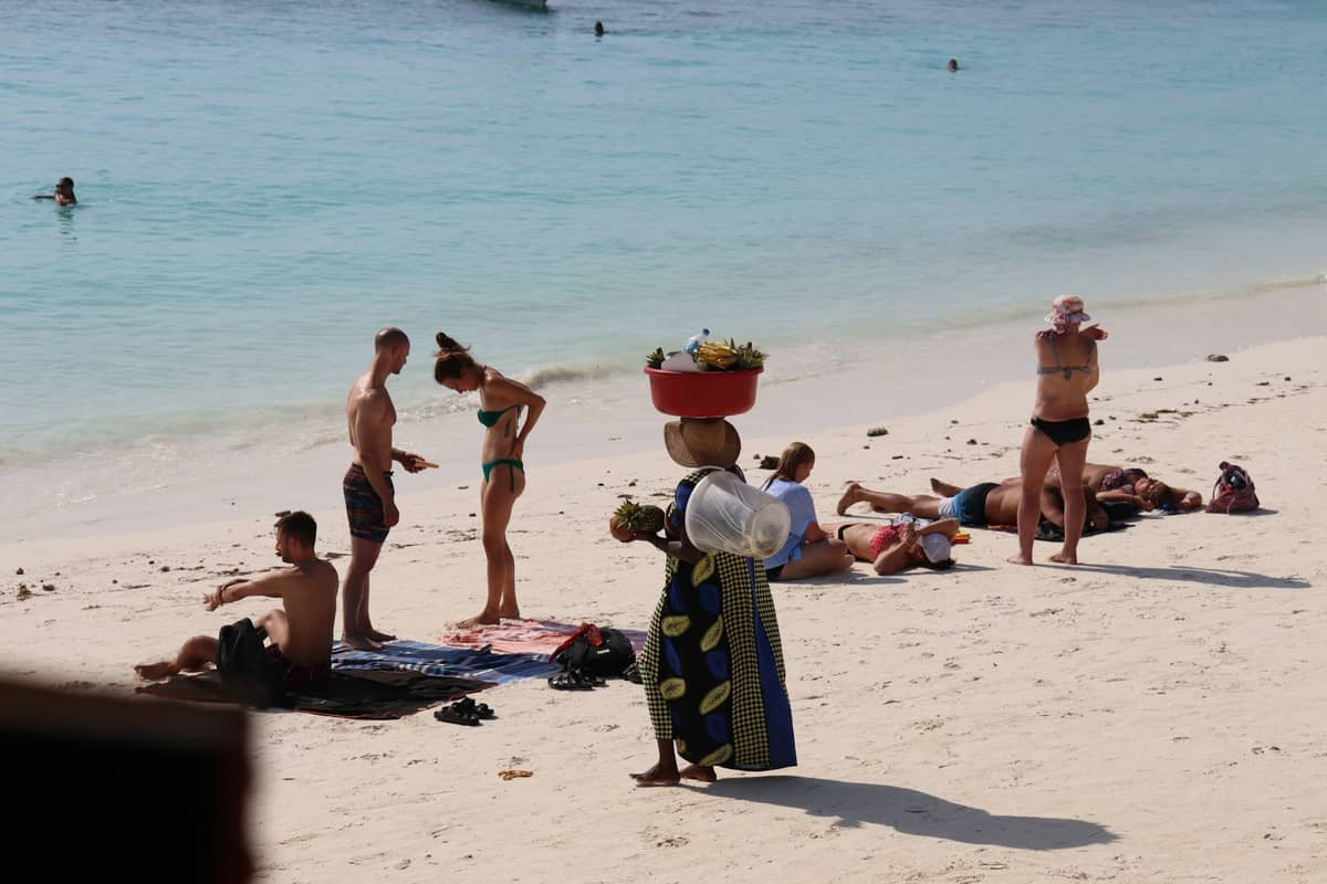 Turistas tomando banho de sol numa praia de areia e uma mulher local carregando mercadorias na cabeça, com o mar ao fundo em Zanzibar.