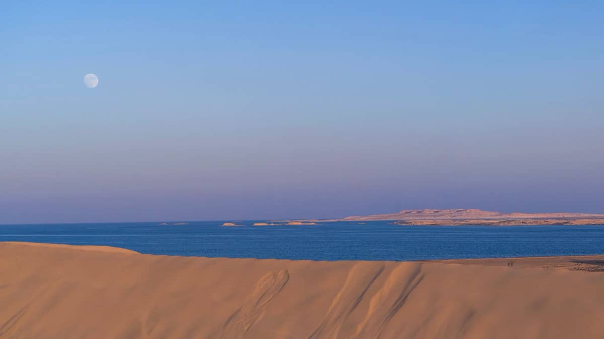 Goldene Sanddünen treffen auf das türkisfarbene Meer unter einem Pastellhimmel mit sichtbarem Mond, was eine Landschaft andeutet, wo die Wüste auf das Meer trifft, in der Nähe von Doha.