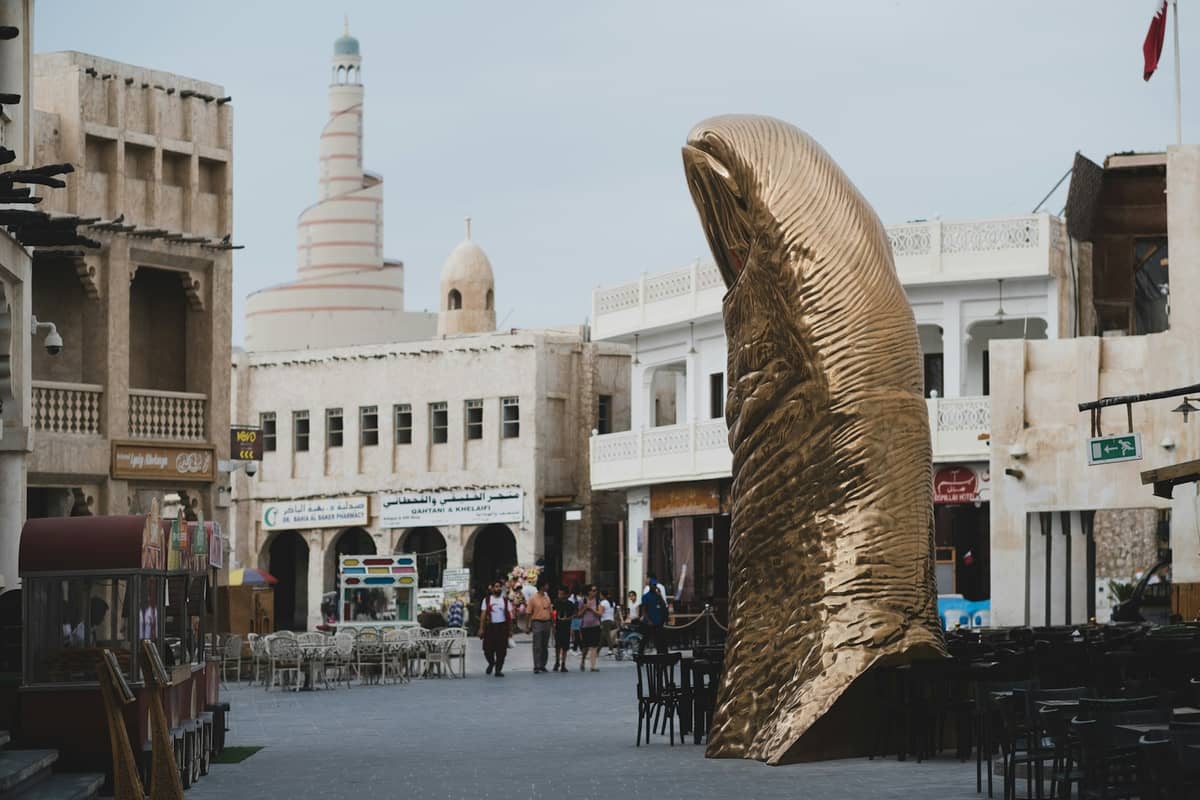 Straßenansicht des Souq Waqif in Doha mit der goldenen Daumen-Skulptur, traditionellen Gebäuden und einem Spiralminarett im Hintergrund.