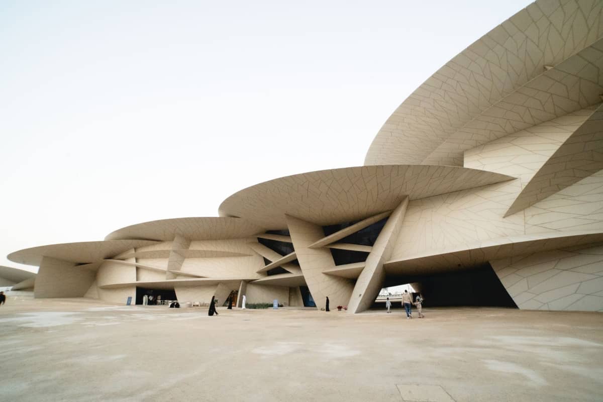 Weitwinkelansicht der dramatischen, ineinandergreifenden Scheibenarchitektur des Nationalmuseums von Qatar mit Besuchern, die auf dem weitläufigen Vorplatz spazieren.