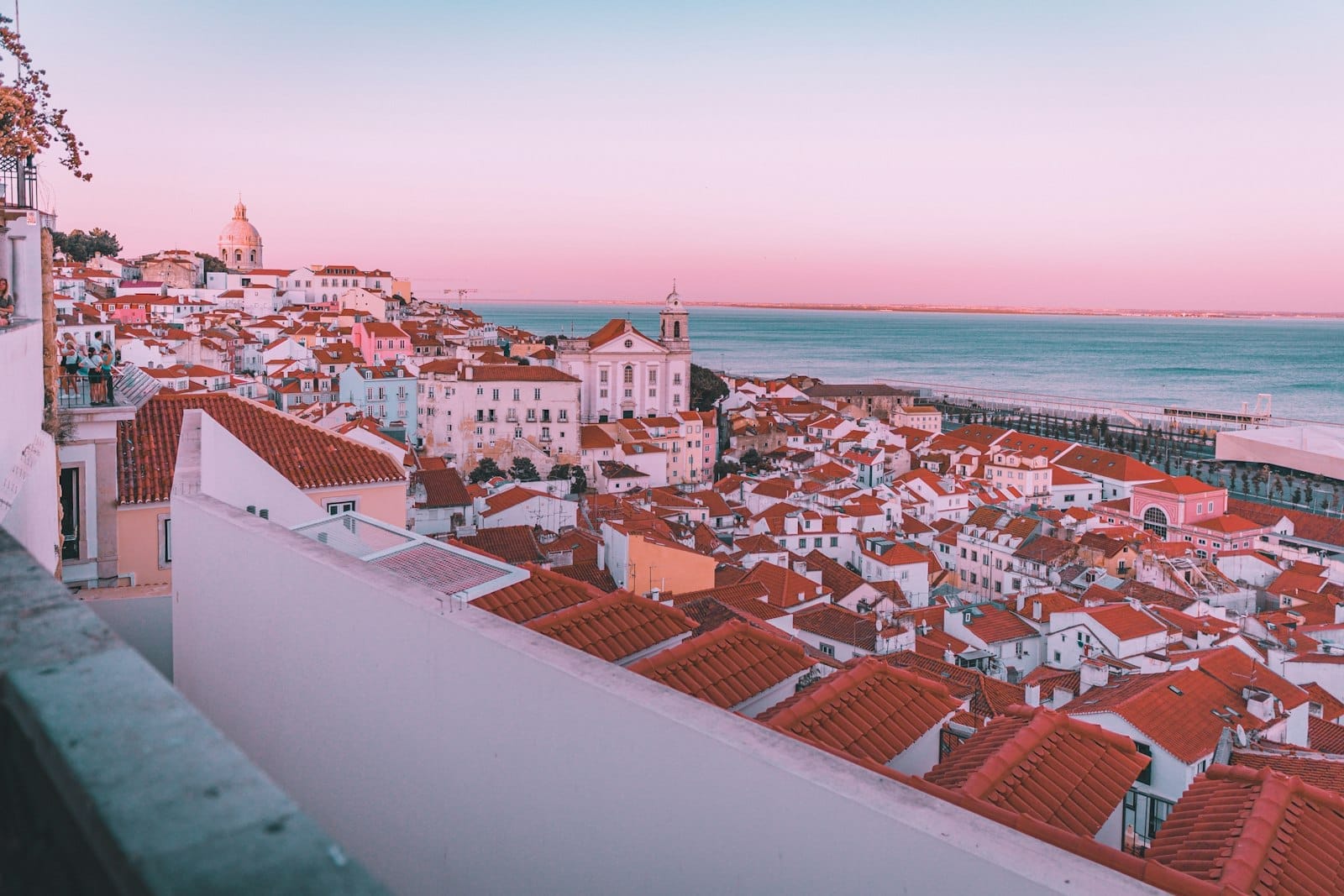 Vista panorâmica deslumbrante dos telhados vermelhos de Lisboa e do Rio Tejo ao amanhecer ou entardecer, com céu rosado e icônicos prédios históricos.