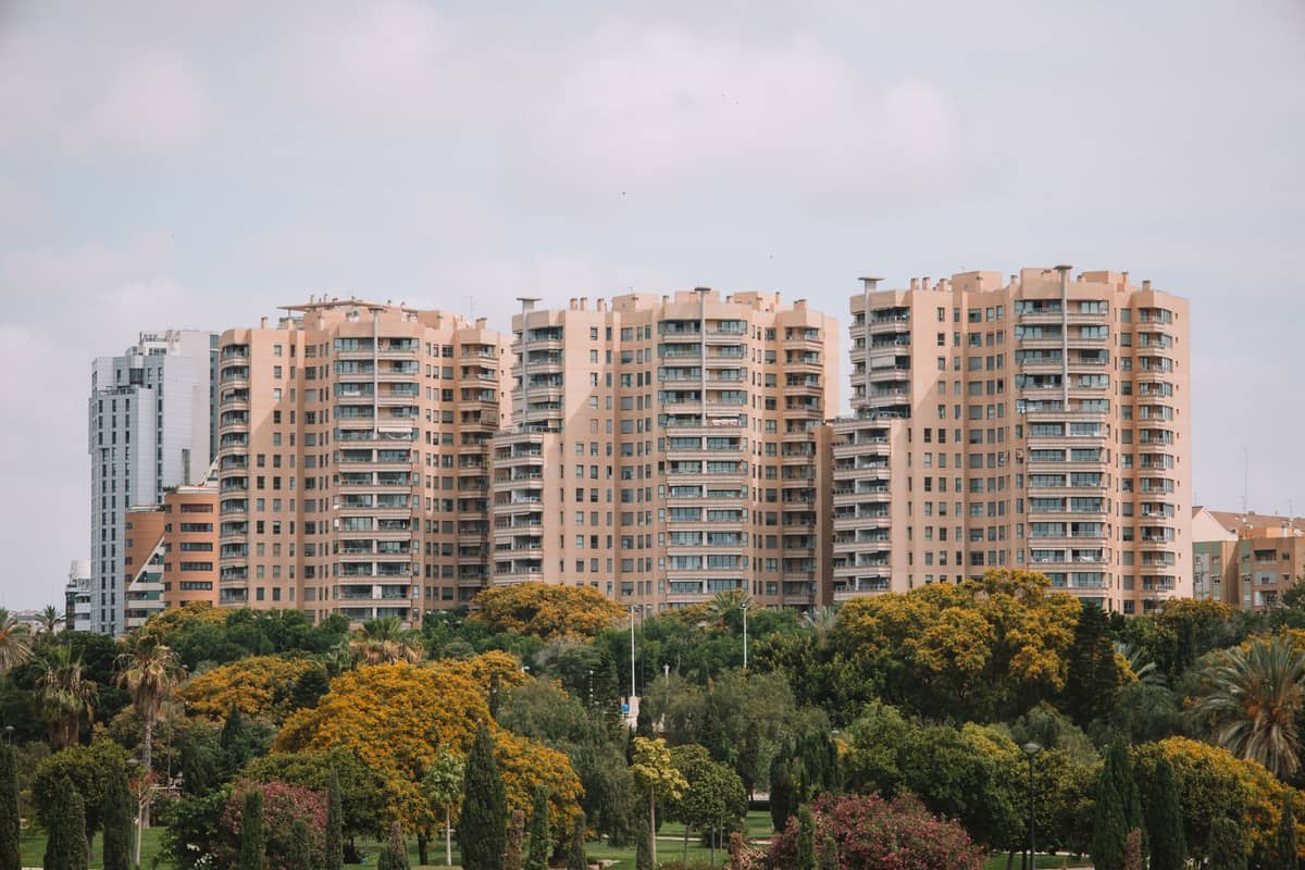 Alti edifici residenziali si innalzano sopra rigogliosi alberi verdi nel Jardín del Turia di Valencia in una giornata luminosa e nuvolosa.
