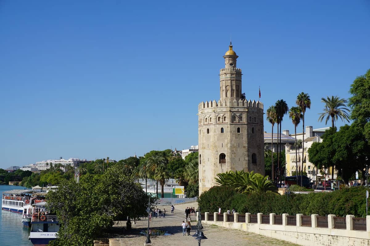 วิวริมแม่น้ำของ Torre del Oro ในเมือง Seville พร้อมต้นปาล์ม ทางเดินเลียบแม่น้ำ และเรือจอดเทียบท่าตามแนว Guadalquivir ภายใต้ท้องฟ้าสีฟ้าแจ่มใส