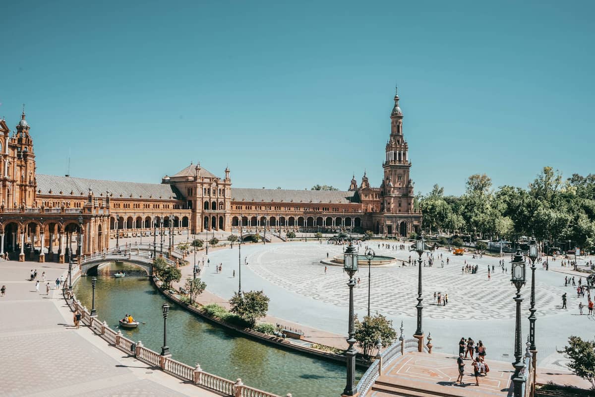 วิวกว้างของ Plaza de España ในเมืองเซบีญา พร้อมคลอง น้ำพุกลาง และอาคารโค้งขนาดใหญ่ภายใต้ท้องฟ้าสีฟ้าแจ่มใส