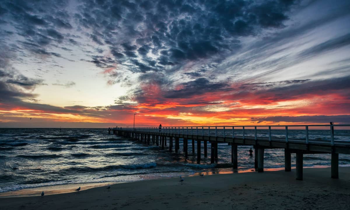 A long wooden jetty extends into the ocean at sunset, with dramatic clouds and vivid orange-red sky over the water.