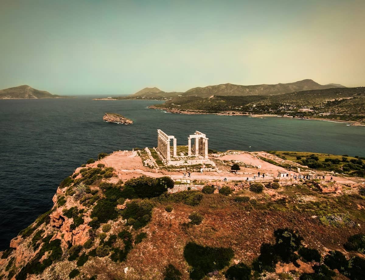 Luftaufnahme des Poseidon-Tempels am Kap Sounion auf einer dramatischen Klippe mit Blick auf das Meer und Bergen im Hintergrund.