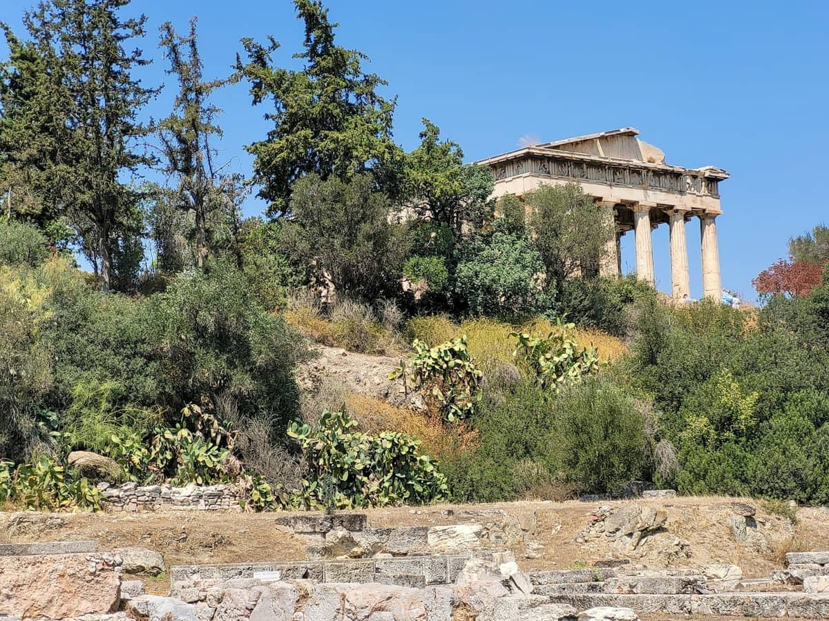 Antiker griechischer Tempel auf einem Hügel, umgeben von Bäumen und Sträuchern unter einem klaren blauen Himmel in Athen.