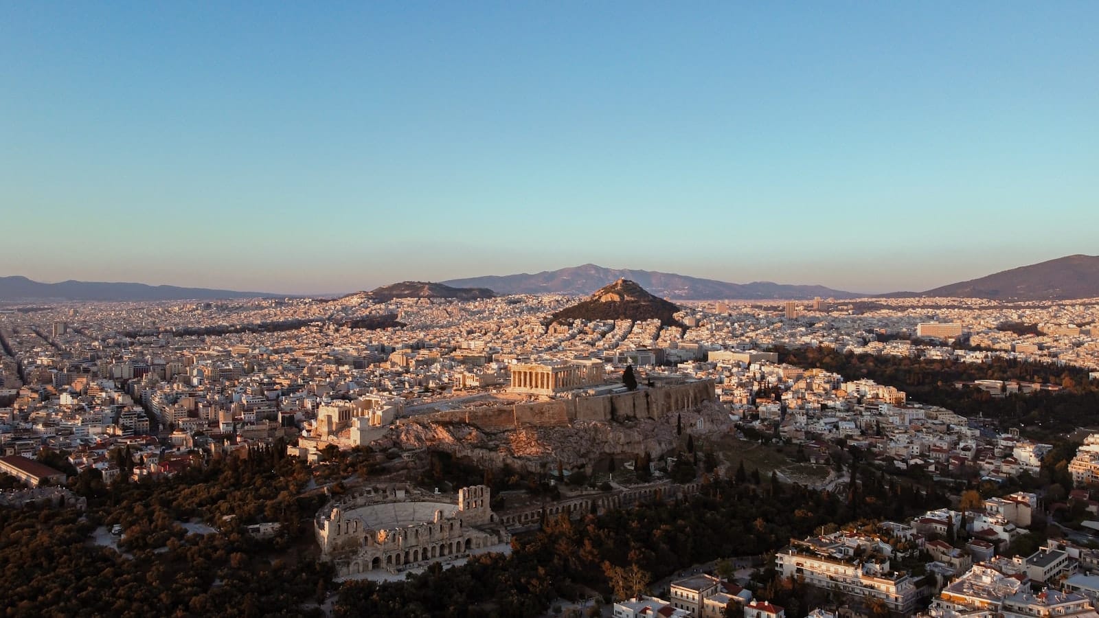 Vista panorâmica de Atenas ao pôr do sol, com a Acrópole, o Partenon, a paisagem urbana e colinas ao fundo sob um céu límpido.