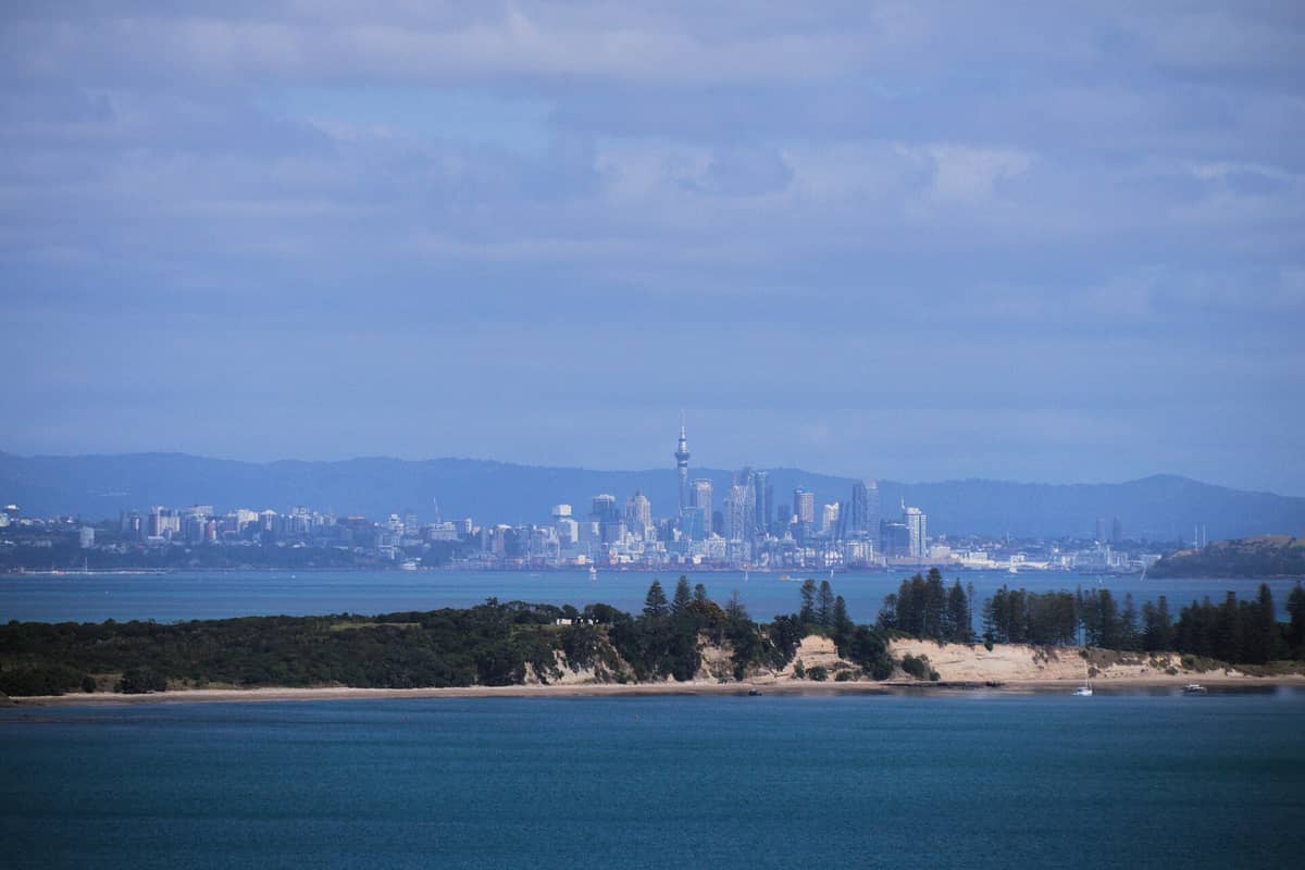 Panorama miasta Auckland widoczna w oddali za wodą, z wyspą i piaszczystym brzegiem na pierwszym planie.