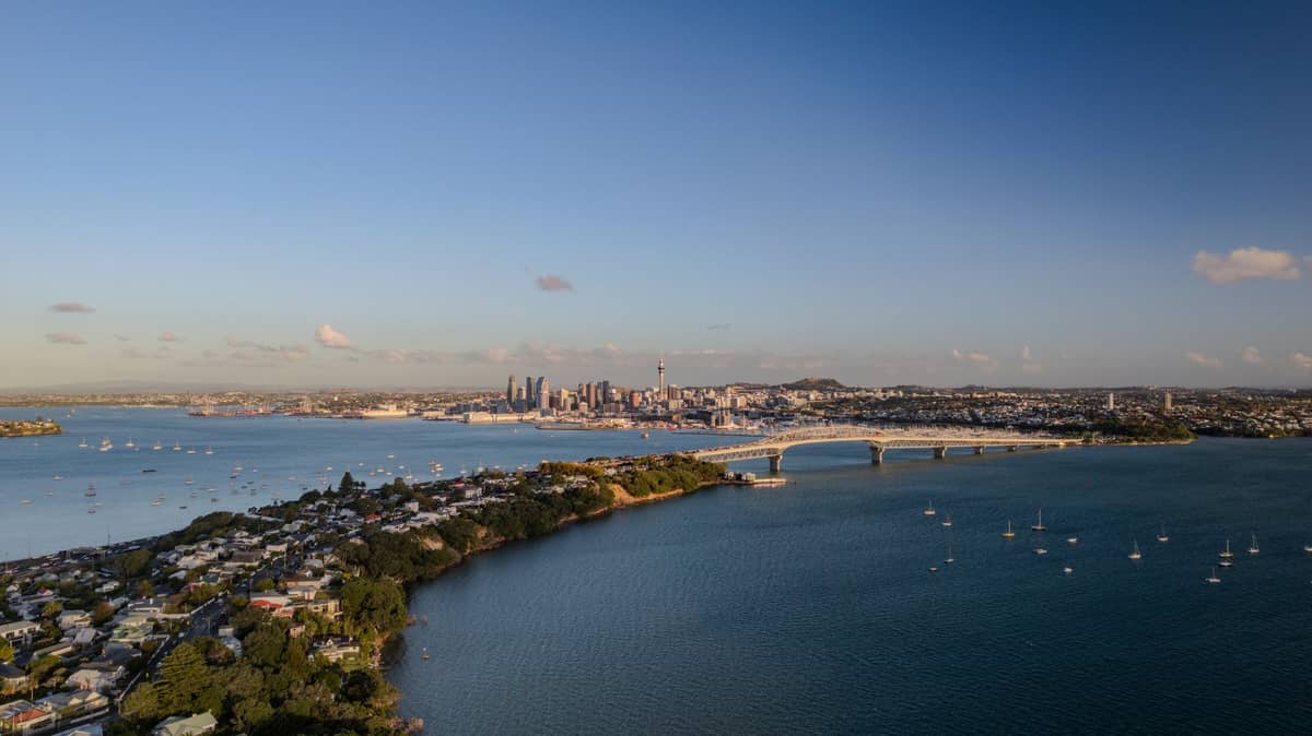 Szeroki widok z lotu ptaka na nabrzeże i panoramę Auckland z Sky Tower, mostem portowym i spokojną niebieską wodą w pogodny dzień.