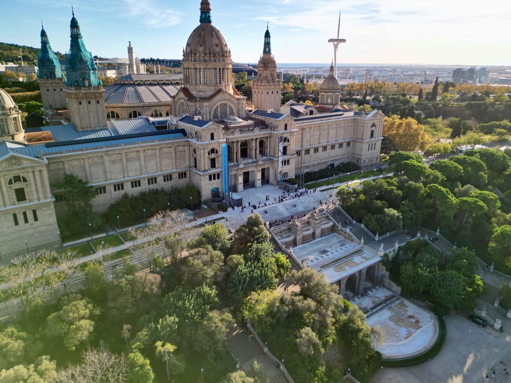 Aerial view of the MNAC palace and Montjuïc hill surrounded by trees and urban landscape in Barcelona, under a clear blue sky.