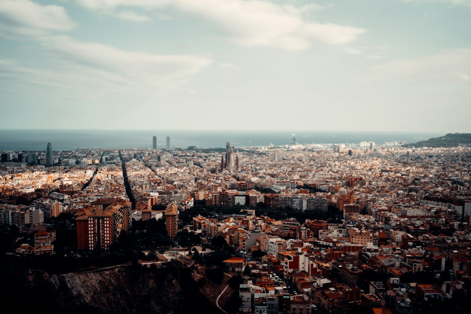 Sweeping panoramic view over Barcelona from a high vantage point with the cityscape, iconic Sagrada Familia, and the Mediterranean Sea under a dramatic sky.