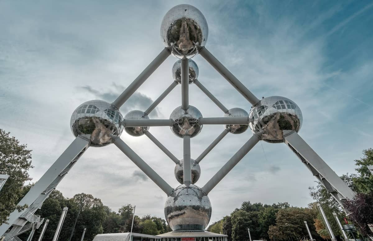 Vista ampla do Atomium em Bruxelas tirada ao nível do solo, mostrando suas esferas prateadas futuristas e a vegetação ao redor sob um céu azul.