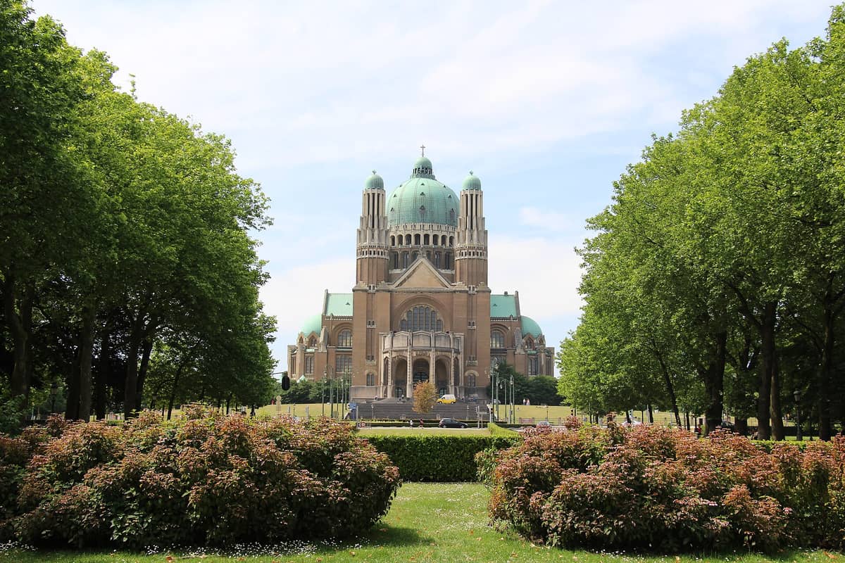 Vista diurna ampla da Basílica Nacional do Sagrado Coração em Koekelberg, emoldurada por árvores e jardins, sob um céu parcialmente nublado.