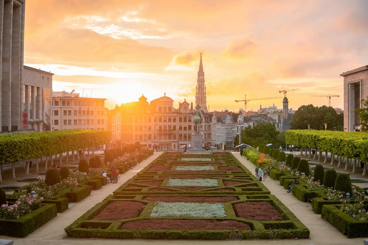 Vista panorâmica do Mont des Arts em Bruxelas ao pôr do sol, com telhados, jardins e a torre da Câmara Municipal ao fundo.
