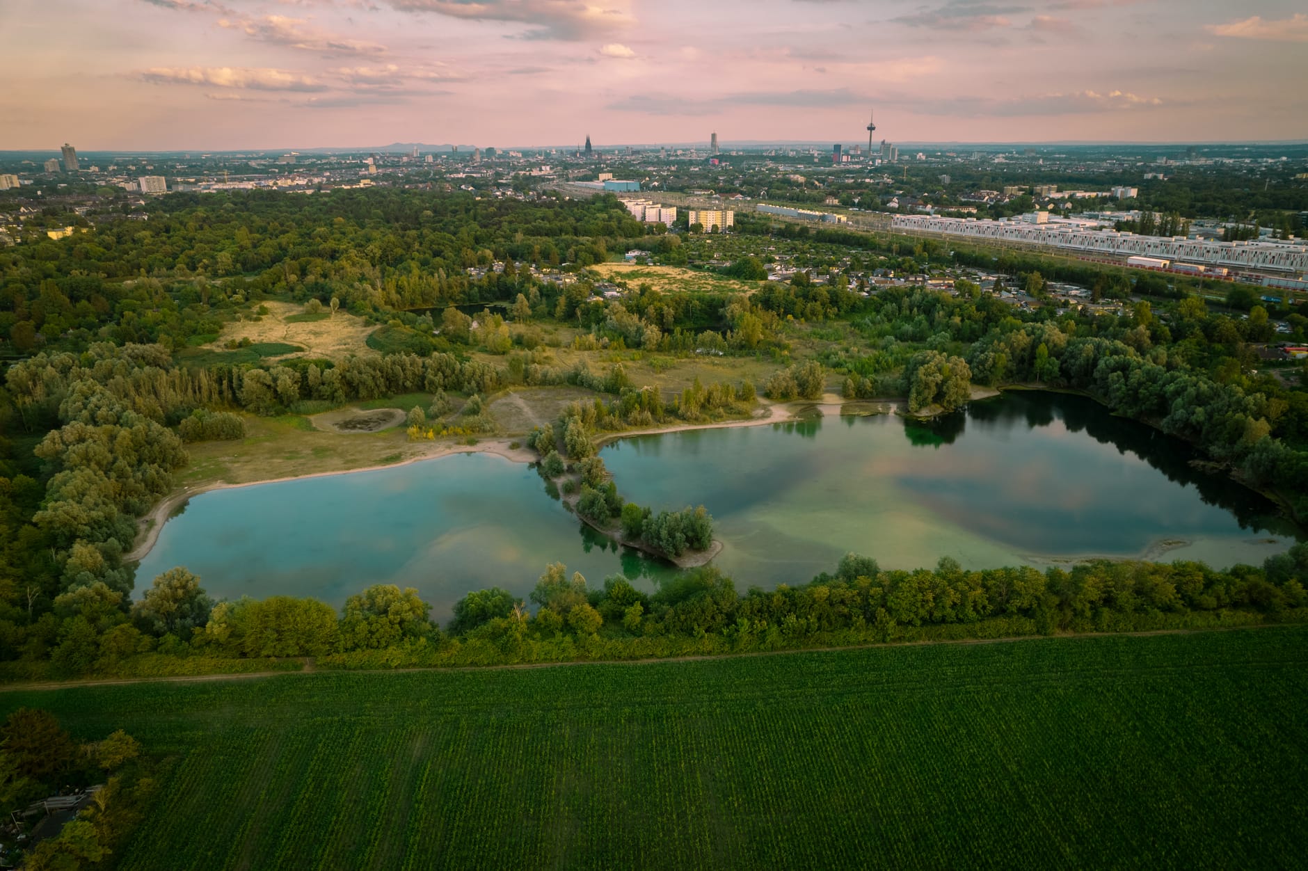 มุมมองทางอากาศของ Cologne แสดงให้เห็นสวนสาธารณะสีเขียวขนาดใหญ่ ทะเลสาบ และเส้นขอบฟ้าของเมืองในพื้นหลังภายใต้ท้องฟ้าหลากสี