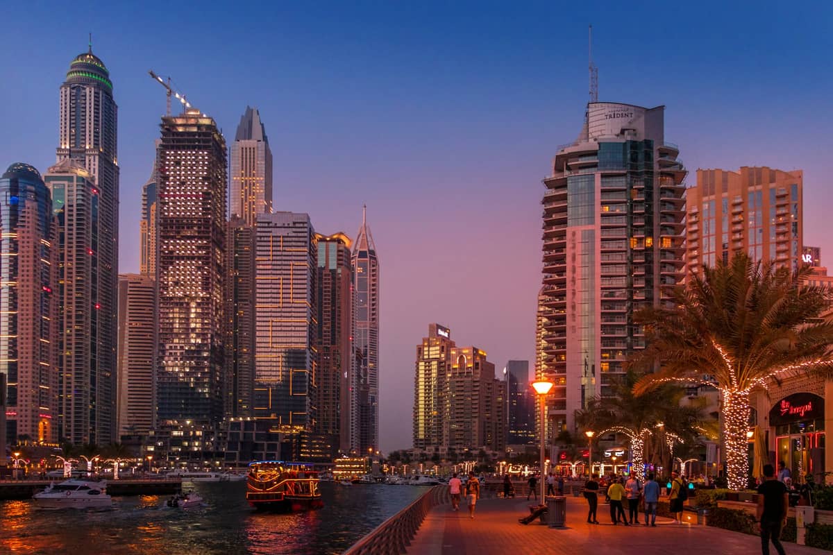 Uferpromenade der Dubai Marina in der Abenddämmerung mit Spaziergängern, beleuchteten Palmen, Booten auf dem Wasser und erleuchteten Wolkenkratzern vor einem lila Himmel.