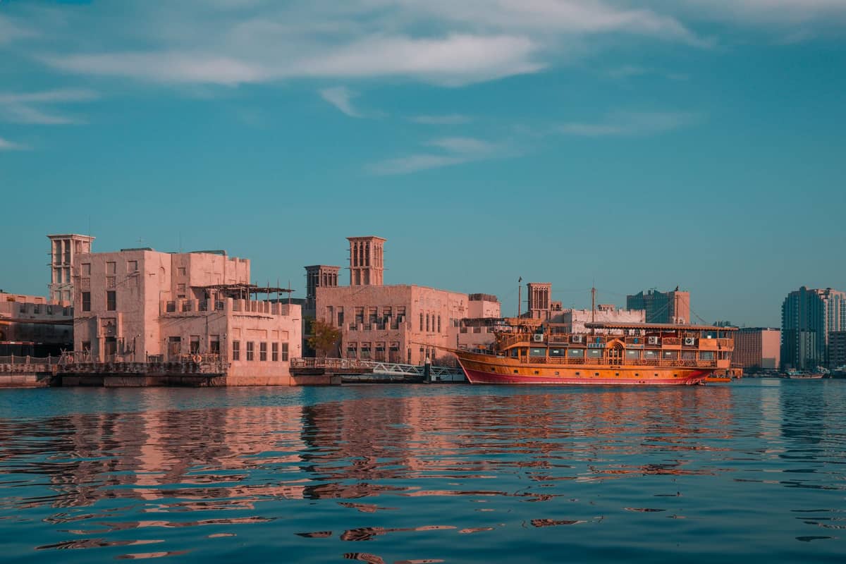 Windtürme und traditionelle Gebäude säumen den Dubai Creek, während ein hölzernes Dau-Boot am Wasser vertäut ist und die Erbe-Architektur des alten Dubais widerspiegelt.