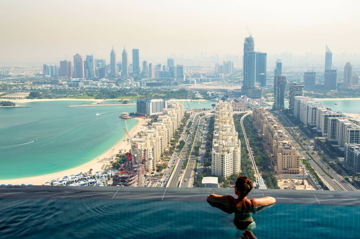 Eine Frau entspannt in einem Infinity-Pool hoch über Dubai mit Blick auf die Palm Jumeirah, die Skyline, Wolkenkratzer und den Strand darunter.