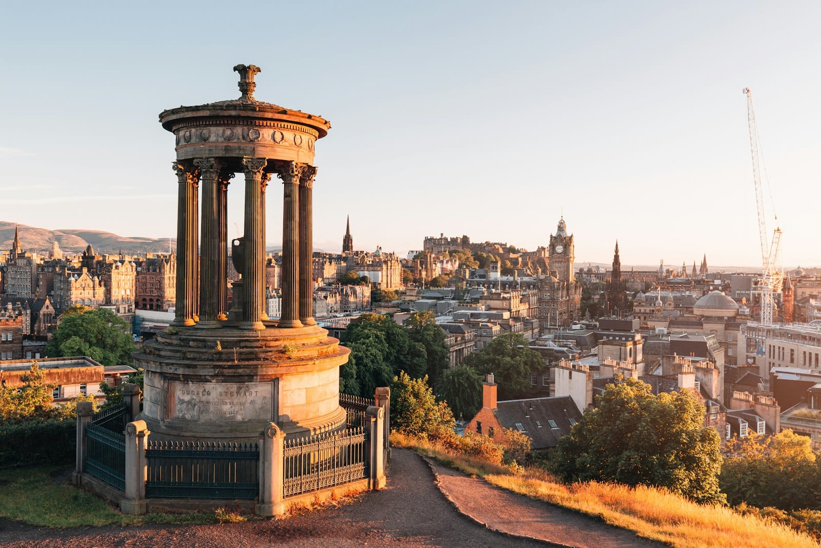 Panoramablick vom Calton Hill über die Skyline von Edinburgh – im Vordergrund das Dugald-Stewart-Monument, im Hintergrund Edinburgh Castle und weitere Wahrzeichen der Stadt bei Sonnenaufgang.