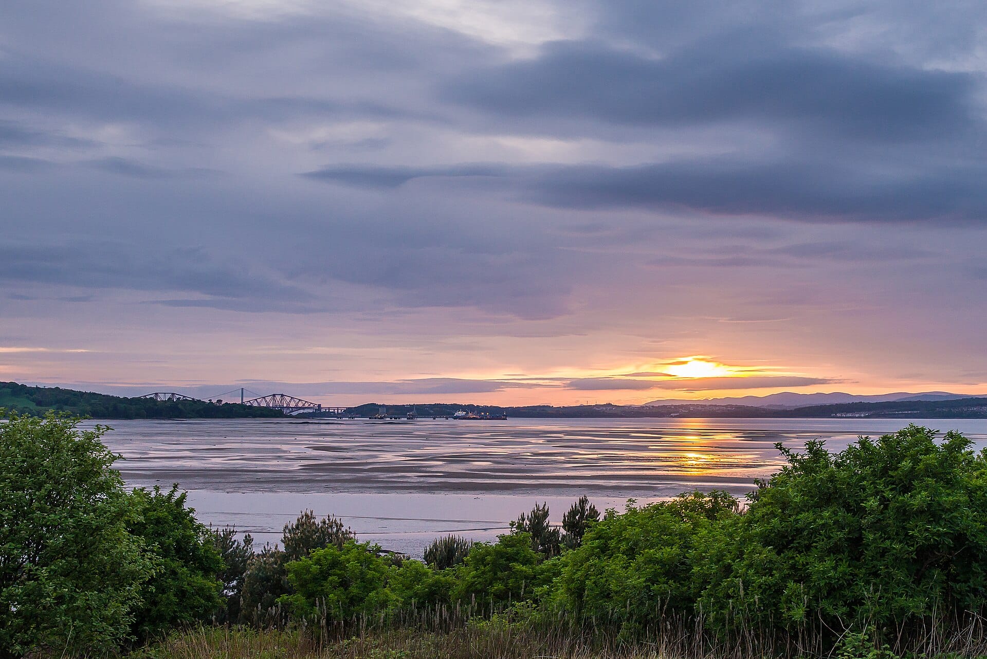 Vue du coucher de soleil sur le Firth of Forth depuis la côte près d'Édimbourg, avec un feuillage vert au premier plan et les Forth Bridges en arrière-plan.