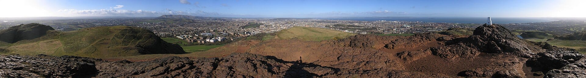 Large panorama depuis le sommet d'Arthur's Seat, montrant des roches volcaniques au premier plan, des collines verdoyantes et une vue expansive sur la ligne d'horizon d'Édimbourg et le littoral sous un ciel bleu.