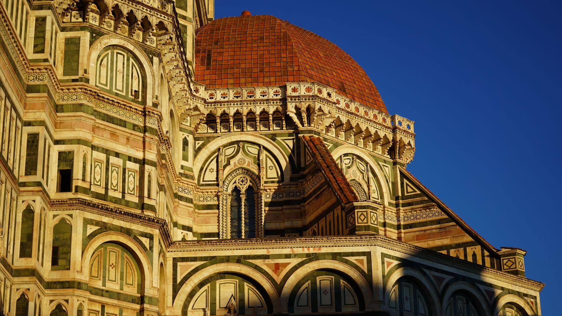 วิวภายนอกของโดมมหาวิหาร Florence และโครงสร้างที่อยู่ติดกันที่สาดส่องด้วยแสงแดดสีทอง แสดงให้เห็นรายละเอียดหินอ่อนที่ประณีตและหลังคากระเบื้องสีแดงตัดกับท้องฟ้าสีน้ำเงิน