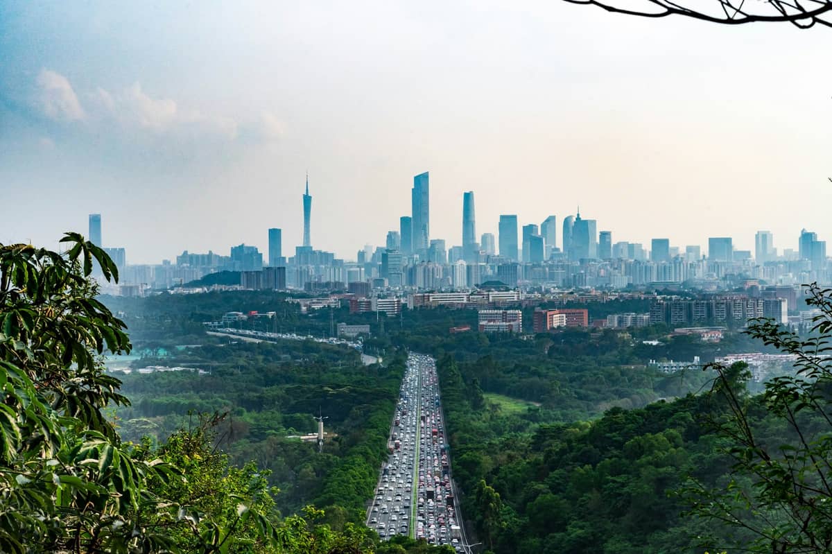 Guangzhou skyline viewed from a green hilltop with Canton Tower and city skyscrapers rising above dense forest and a busy highway leading into the city.