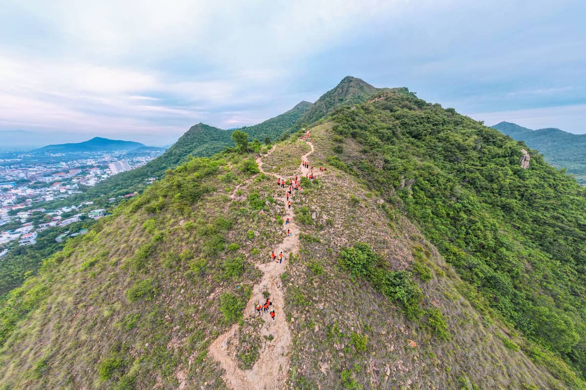 A high aerial view of hikers ascending a green, rounded mountain ridge with cityscape visible in the background under a cloudy sky.