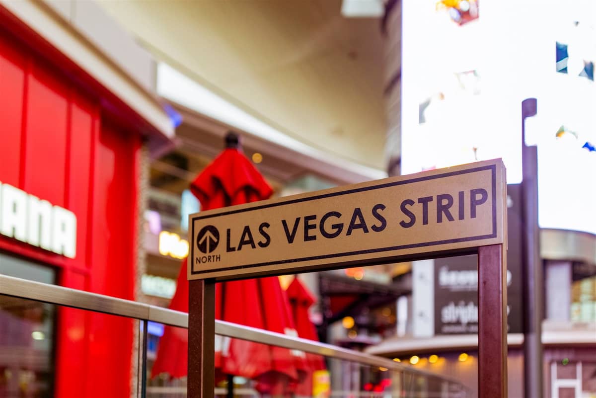 ป้ายที่มีข้อความว่า 'Las Vegas Strip' พร้อมแสงไฟ อาคาร และหน้าร้านที่เบลอในพื้นหลังระดับถนนในช่วงกลางวัน