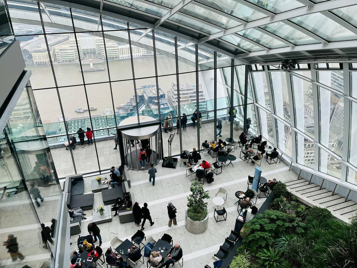 Vista interior de um grande jardim de cobertura envidraçado em Londres com vegetação exuberante, pessoas apreciando as vistas da cidade e o Tâmisa através de janelas panorâmicas.