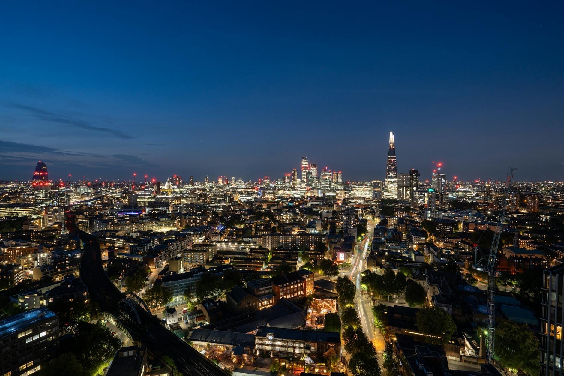 Skyline de Londres ao entardecer com arranha-céus iluminados, The Shard e outros edifícios sob um céu azul profundo, mostrando vistas panorâmicas e as luzes da cidade.