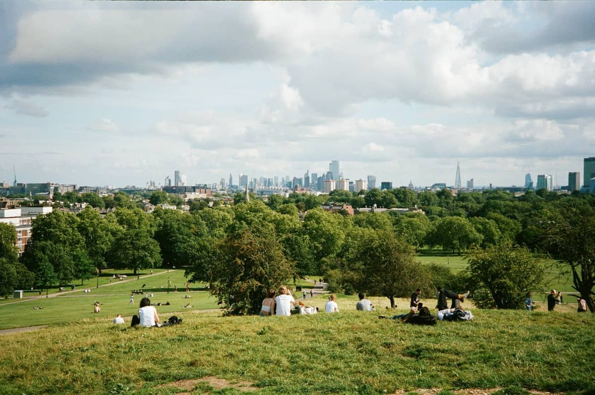 Vista panorâmica do topo de uma colina gramada em um parque de Londres, com pessoas relaxando e o horizonte da cidade e vegetação ao fundo.