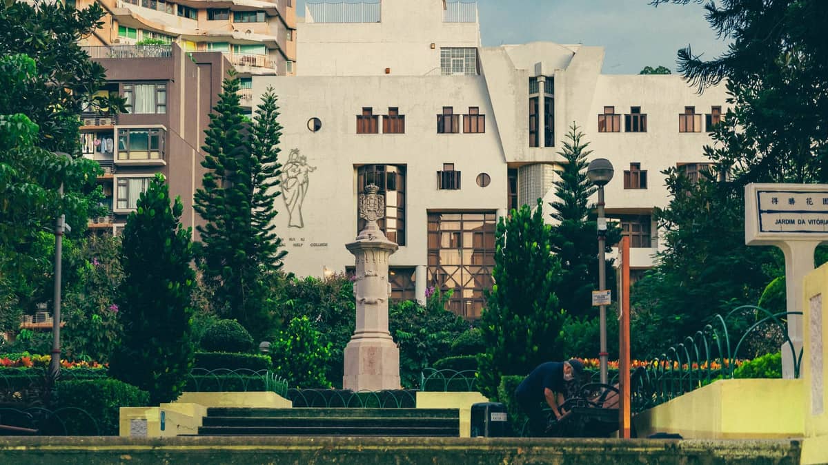 Blick auf ein Art-déco-Gebäude mit einzigartigen Fenstern, umrahmt von angelegten Gärten und hohen Apartmentgebäuden, aufgenommen aus einem grünen Stadtpark in Macau.