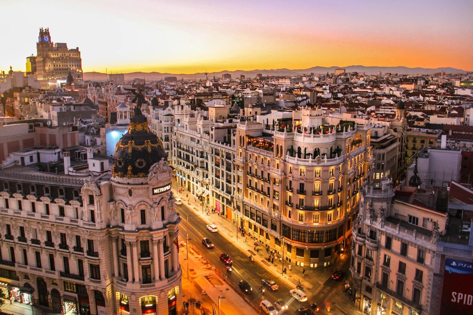 Vista aérea impressionante da Gran Vía de Madri e do Edifício Metrópolis ao pôr do sol, com as luzes quentes da cidade e um céu colorido destacando o famoso skyline madrileno.