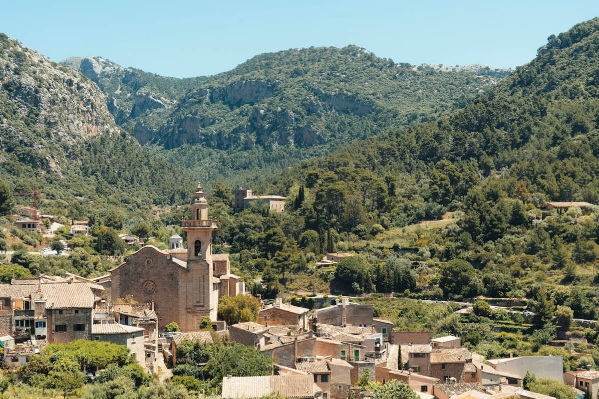 Villaggio in cima a una collina a Mallorca circondato da montagne verdi e foresta, con una chiesa prominente al centro sotto un cielo azzurro luminoso.