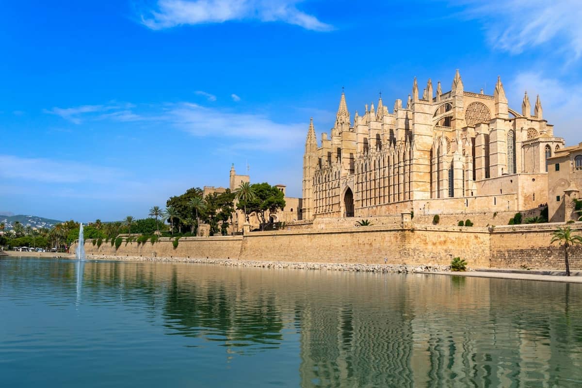 Vista sul lungomare della Cattedrale di Palma e il suo riflesso sul lago, con cielo azzurro e palme in primo piano.