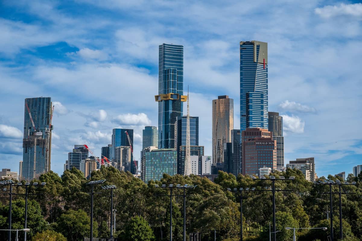 Widok na panoramę Melbourne z nowoczesnymi wieżowcami, w tym Eureka Tower, wznoszącymi się ponad zielenią pod błękitnym niebem.