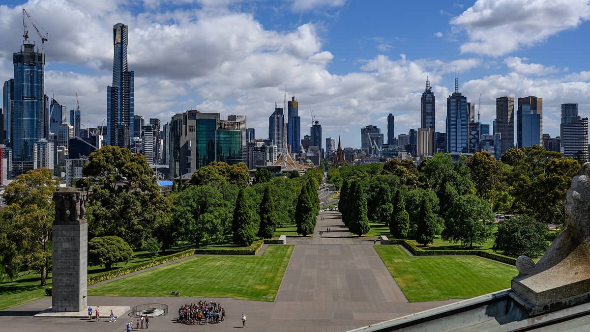 Rozległy widok ze Shrine of Remembrance na zielone ogrody i trawniki Melbourne ze skyline miasta w tle pod częściowo zachmurzonym niebem.