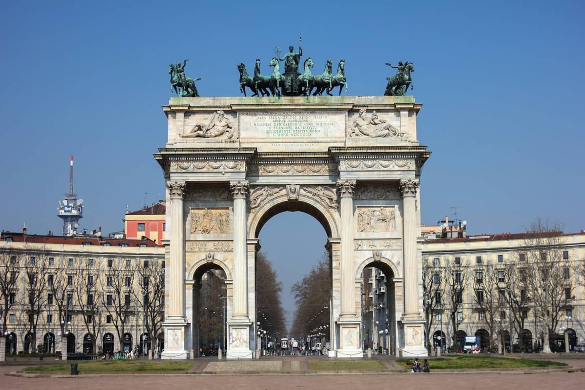 Arco della Pace w Mediolanie widziany z szerokiego, otwartego placu z wyraźnymi osiami widokowymi i otaczającymi budynkami pod błękitnym niebem