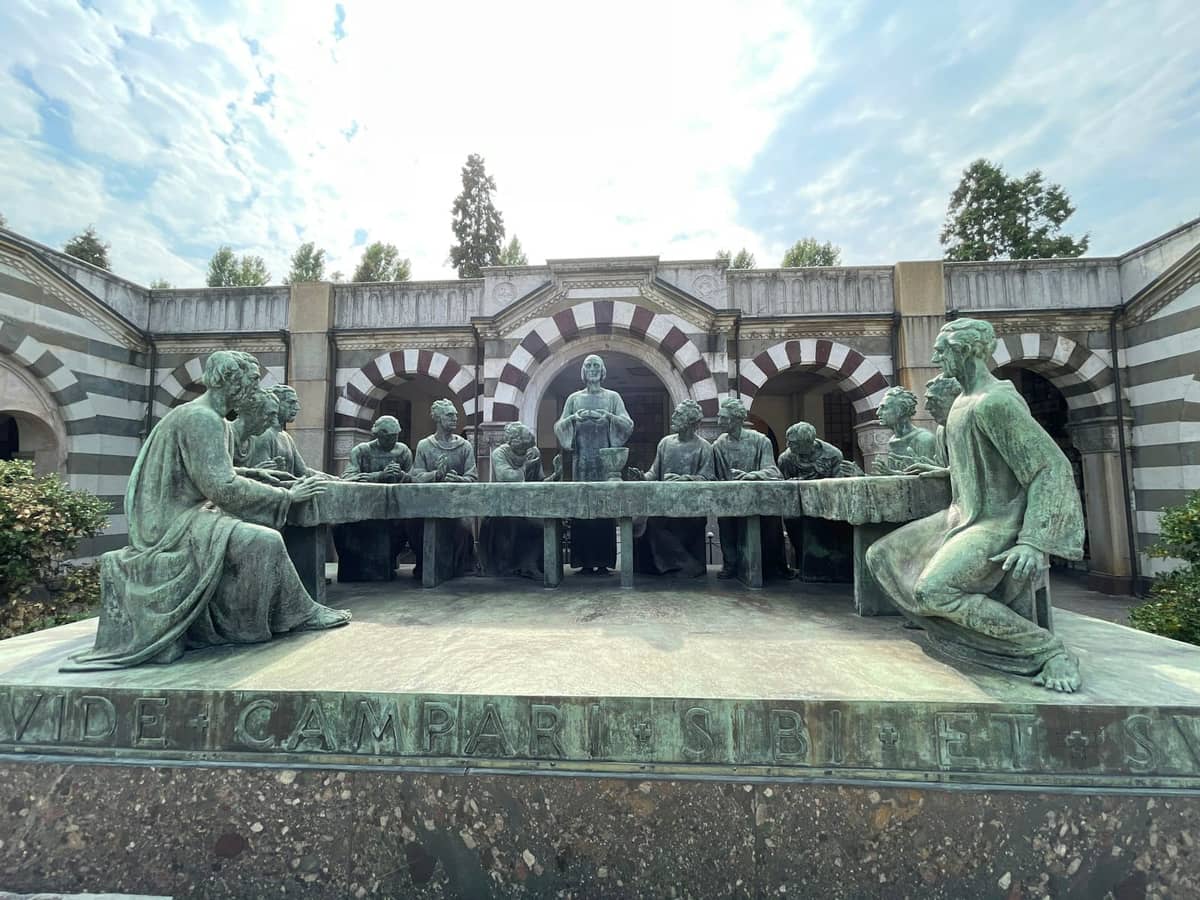 Brązowa rzeźba Ostatniej Wieczerzy na dziedzińcu Cimitero Monumentale w Mediolanie, ze szczegółowymi postaciami i pasiastymi łukami widocznymi pod częściowo zachmurzonym niebem.