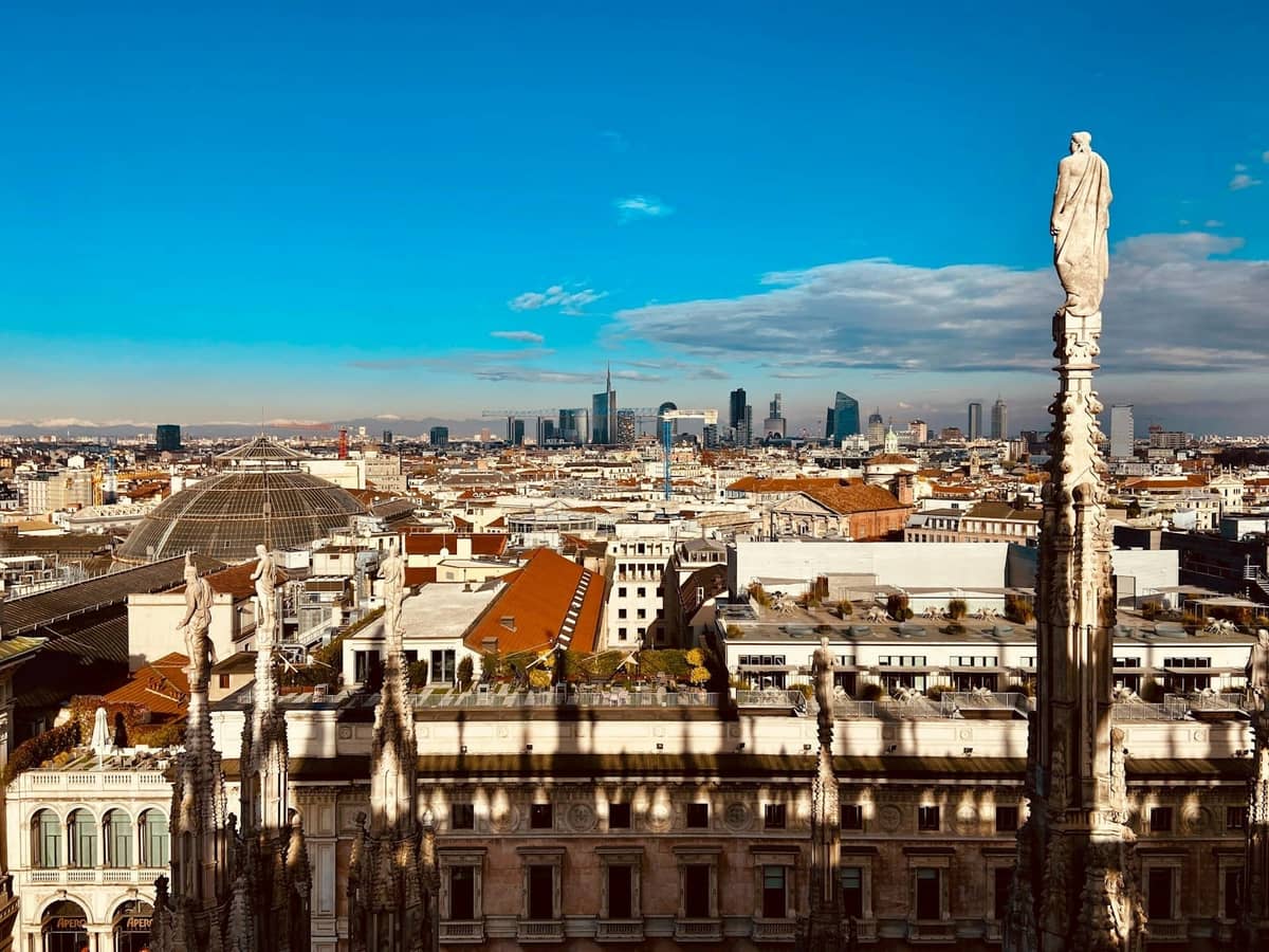 Panoramiczny widok z dachu Katedry w Mediolanie przedstawiający marmurowe iglice, posągi, budynki miejskie i odległą panoramę pod błękitnym niebem.