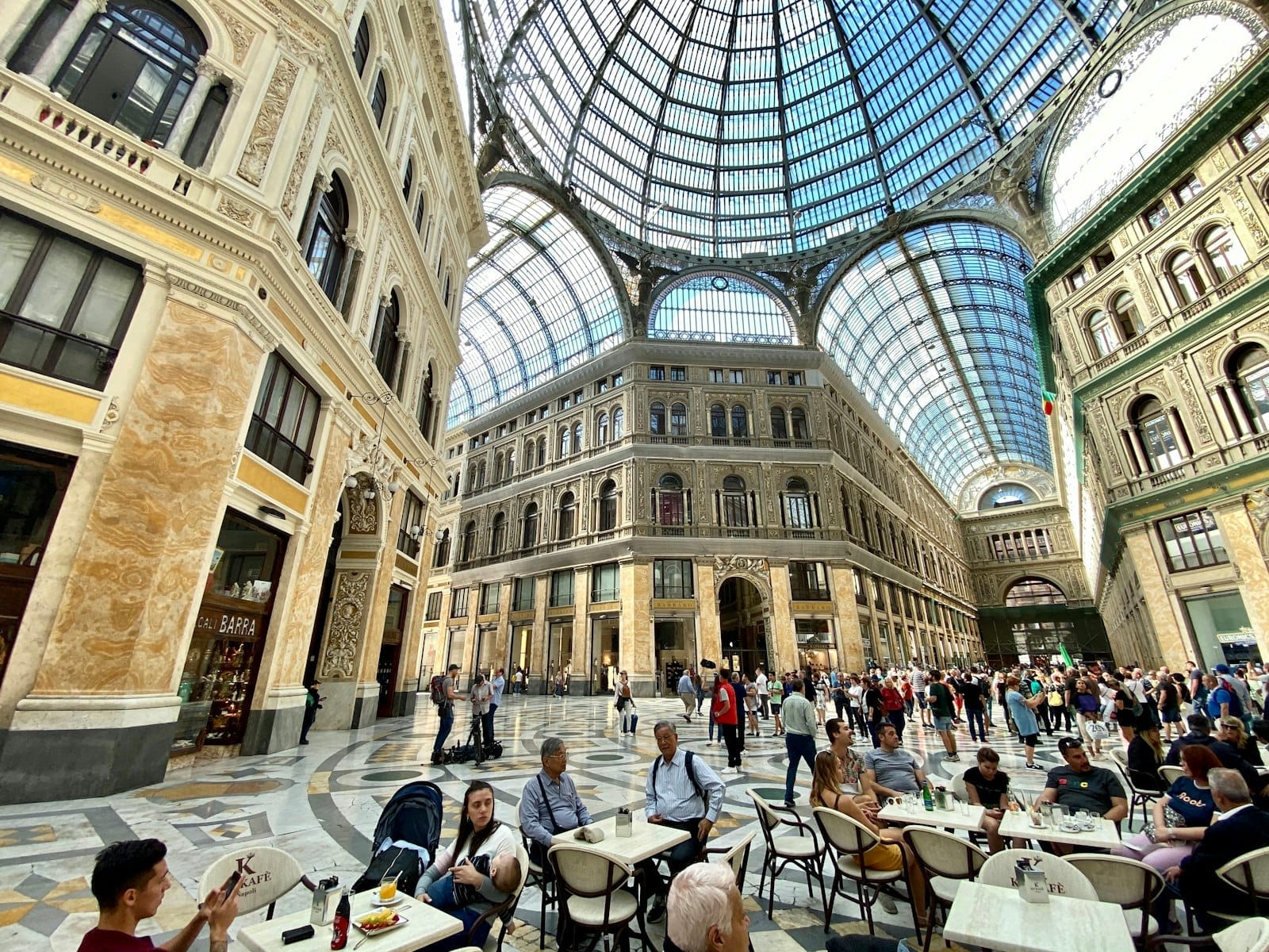 Pessoas jantando e caminhando sob a ornamentada cúpula de vidro da Galleria Umberto I, um grandioso interior arquitetônico em Nápoles, Itália.